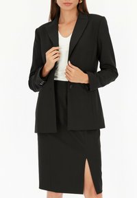 Czarny zestaw żakietu i spódnicy ołówkowej. Żakiet ma dopasowany krój, klapy w kształcie notcha oraz dwa guziki; spódnica ma boczne rozcięcie i gładką fakturę.