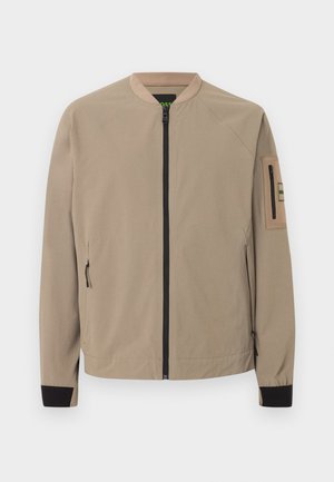 Μπεζ μπουφάν bomber με μαύρο φερμουάρ μπροστά, φερμουάρ στα πλαϊνά τσέπες, μαύρα μανίκια στους καρπούς και τσέπη με φερμουάρ στο αριστερό μανίκι.