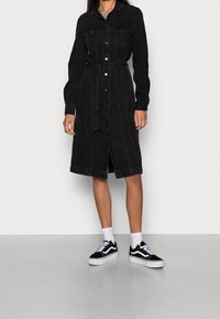 Nainen, joka pitää yllään mustaa nappilista denimimekkoa, jossa on vyötärösolki, yhdistettynä valkoisiin sukkiin ja mustavalkoisiin tennareihin, seisoo valkoista seinää vasten.