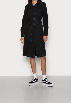 Nainen, joka pitää yllään mustaa nappilista denimimekkoa, jossa on vyötärösolki, yhdistettynä valkoisiin sukkiin ja mustavalkoisiin tennareihin, seisoo valkoista seinää vasten.