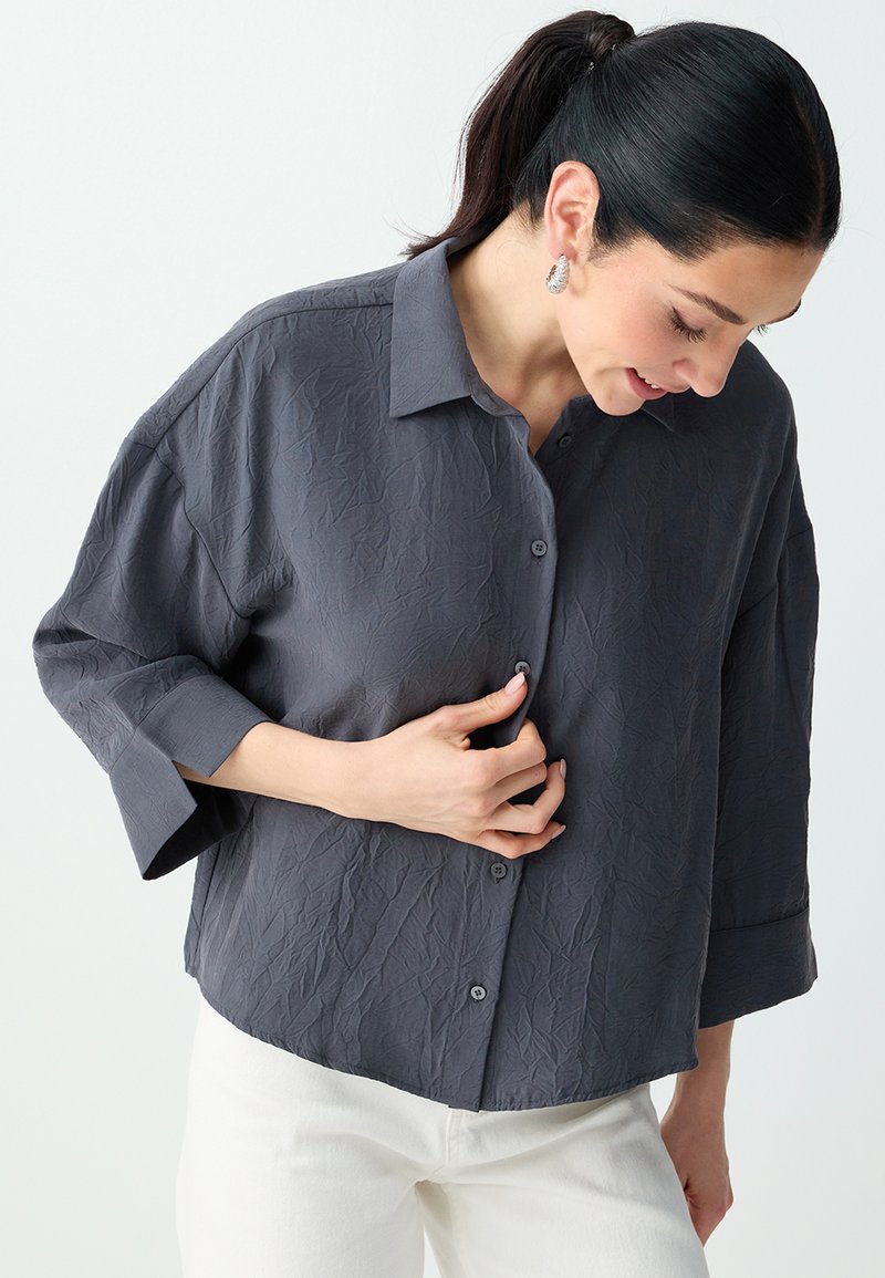 Femme aux cheveux foncés en queue de cheval, portant des boucles d'oreilles argentées, une chemise boutonnée gris foncé froissée et un pantalon blanc, regardant vers le bas et boutonnant un bouton.