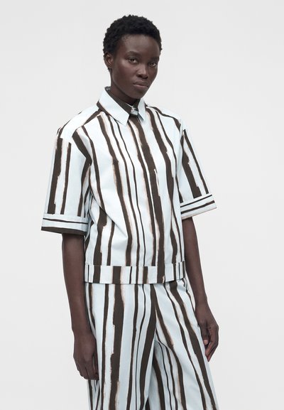 Γυναίκα φορεί χαλαρό πουκάμισο με κοντά μανίκια και παντελόνι με κάθετες μαύρες και λευκές ρίγες, με απλό φόντο.