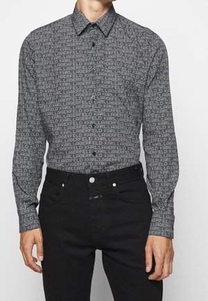 Homme portant une chemise noire à boutons avec le texte blanc répété "Karl Lagerfeld", associée à un jean noir, debout devant un fond uni.