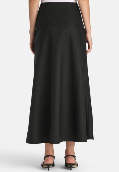 Kvinde iført en lang, højtaljet sort nederdel kombineret med sorte sandaletter med stropper og hæle, set bagfra.