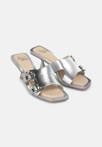 Mule argentati con tacco alto e punta quadrata, caratterizzati da una fascia ampia e due fibbie metalliche circolari, texture in pelle liscia e soletta beige.