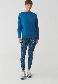 Kék pulóver széles nyakkivágással, kék mintás leggings-szel és világoskék sportcipővel párosítva. Sima anyag, testhezálló kialakítás és kontrasztos minták.
