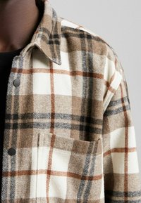 Brązowa, biała i czarna w kratę wełniana kurtka z czarnymi zatrzaskami i kieszenią na piersi noszona na czarnej koszuli na ciemnej skórze.