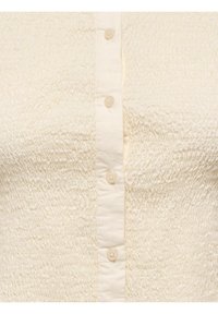 Gros plan d’un tissu couleur crème avec une texture smockée sur les côtés et une patte de boutonnage centrale avec quatre petits boutons ronds.