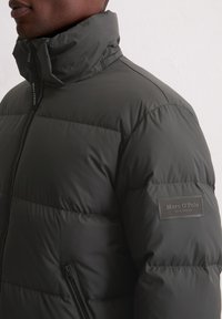 Mężczyzna w ciemnoszarej pikowanej kurtce puchowej z wysokim kołnierzem i zamykaną kieszenią, etykieta Marc O'Polo na rękawie oraz suwakiem.