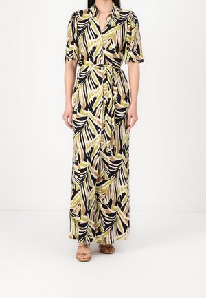 Woman wearing a long, short-sleeved dress with green, black, beige abstract leaf patterns and a tied waist belt, paired with tan sandals.