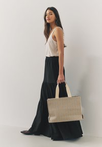 Torba na ramię z pionowymi czarno-beżowymi paskami, wykonana z tkanego materiału. Modelka ubrana w biały top na ramiączkach i czarną wielowarstwową spódnicę, trzyma torbę.