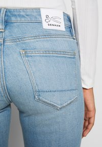 Jean en denim bleu clair avec taille haute, poches arrière et une étiquette avec un logo en forme de ciseaux. Texture lisse et détails de couture classiques.