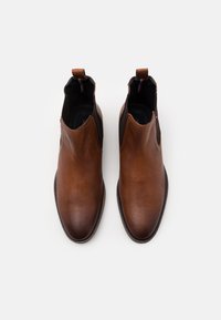 Botas de cuero marrón hasta el tobillo con paneles laterales elásticos, puntera redondeada y asa de tiro. Superficie texturizada con una suela oscura contrastante.