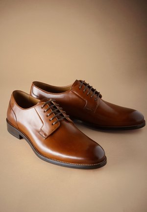 Chaussures habillées pour hommes en cuir marron poli avec semelles noires et lacets marron foncé sur fond beige.