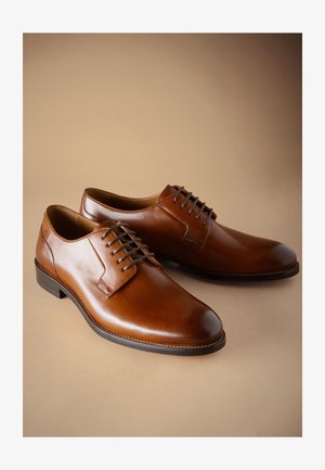 Chaussures habillées pour hommes en cuir marron poli avec semelles noires et lacets marron foncé sur fond beige.