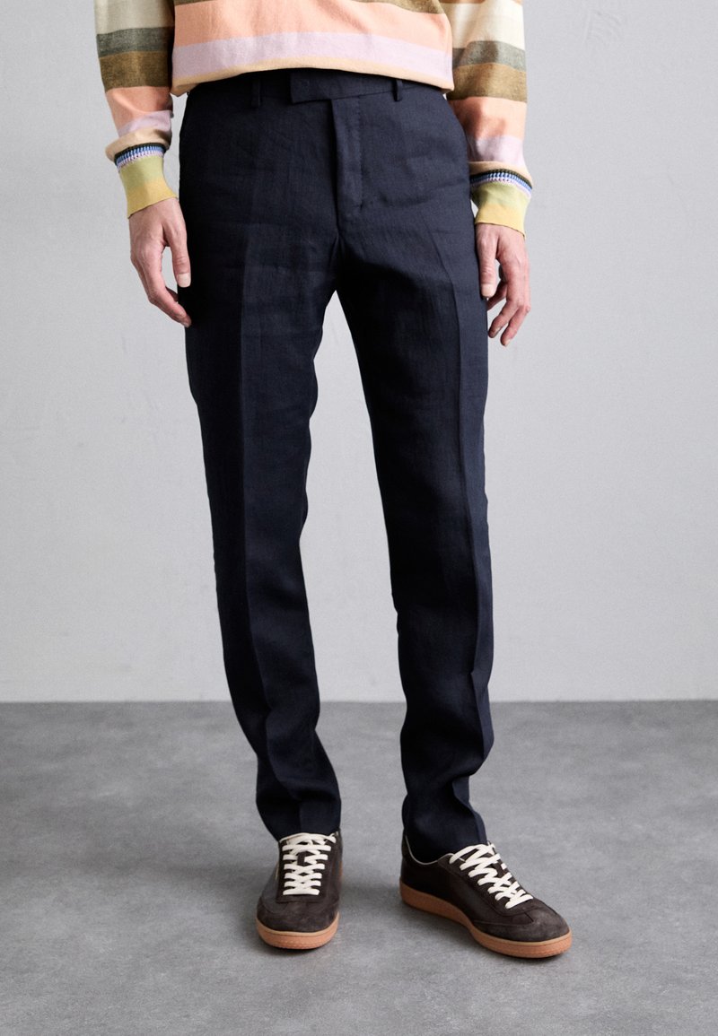 Calças marinhas em tecido leve apresentam um corte de perna reta e uma textura subtil. Combinadas com sapatilhas de camurça escuras e um suéter listrado multicolorido.