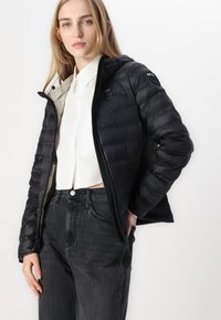 Černá péřová bunda s kapucí, která má hladký povrch, zip a lehký design. Nositá přes bílé tričko a tmavé džíny.