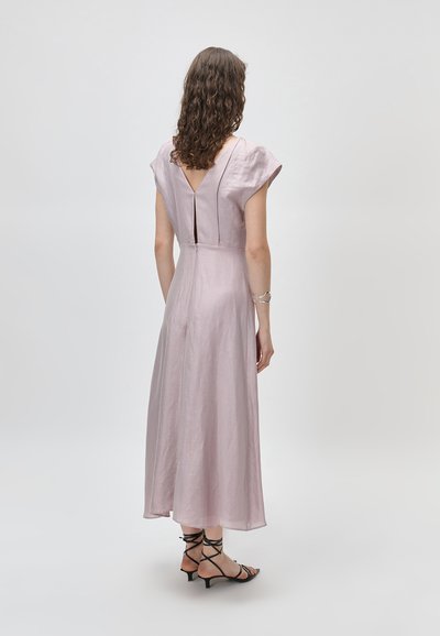 Γυναίκα με καστανά σγουρά μαλλιά φοράει ένα μωβ μεσαίο φόρεμα με κοντά μανίκια και μαύρα σανδάλια με λουράκια και χαμηλό τακούνι, στέκεται σε απλό φόντο.