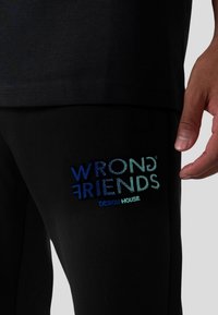 Černé kalhoty s ribbed texturou, které mají barevně vyšívané logo "WRONG FRIENDS DESIGN HOUSE" na levém stehně.