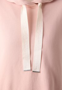 Gros plan d'un sweat à capuche rose pâle avec des cordons blancs présentant un nom de marque subtilement embossé et des embouts métalliques aux extrémités.