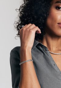 Srebrna pleciona bransoletka na nadgarstku, noszona z ciemną koszulą zapinaną na guziki. Tekstura wzoru z gładkim, wypolerowanym wykończeniem.