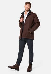 Bruine fleecejas met een zwarte kraag, knopen aan de voorkant en zakken; gedragen over een wit shirt en grijze broek met bruin leren laarzen.