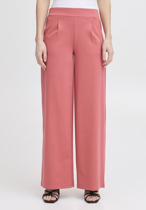 Pantaloni a gamba larga rosa pallido con una texture liscia, dotati di pieghe frontali e una vita alta. Abbinati a sandali neri con cinturini.