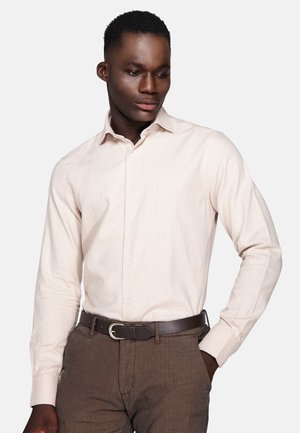 Man wearing a light beige button-up shirt, brown pants with a belt, standing with one hand in pocket, looking down to his left.