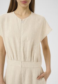 Combinaison en lin beige à manches courtes, avec un col rond et une fermeture éclair à l'avant. Présente une taille froncée et des poches latérales.