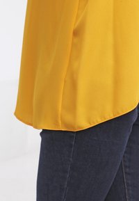 Žlutá blůza s hladkým, lehkým materiálem, zobrazující kulatý lem a jemné drapování, v kombinaci s tmavými džíny.