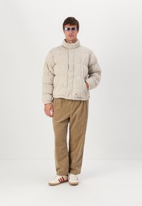 Puffet lys beige jakke med høj krave og quiltet mønster, sammen med brede tan farvede corduroybukser og afslappede hvide sneakers med røde striber.