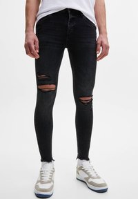Hombre con pantalones ajustados desgastados negros y dobladillos deshilachados, calcetines blancos, zapatillas blancas y beige, y una camiseta blanca frente a un fondo liso.