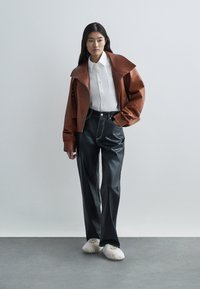 Bruine leren jas met brede kraag, witte button-up shirt, zwarte leren broek en witte pluizige slippers; slanke, moderne silhouet.