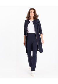 Marineblauwe lange jas met ritssluiting, volledige mouwen, gecombineerd met bijpassende slim-fit broek. Een witte t-shirt en witte sneakers maken de outfit af.