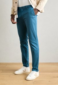 Tyrkysové slim-fit nohavice vyrobené z ľahkého materiálu, s pásom na gombík a bočnými vreckami, v kombinácii s bielymi teniskami.
