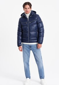 Granatowa kurtka puchowa z kapturem, z zamykanymi na zamek kieszeniami na piersi i błyszczącym wykończeniem, w połączeniu z jasnoniebieskimi dżinsami i białymi trampkami.