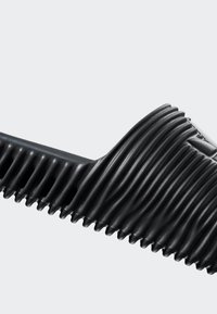 Svart formgjuten slidesandal med en vågformad ribbad textur, öppen tå-design och en platt sula. Tillverkad av flexibelt syntetmaterial.