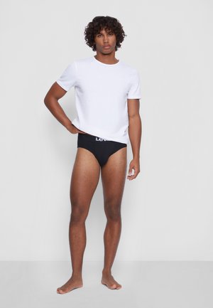 Weißes Baumwoll-T-Shirt mit kurzen Ärmeln, schwarze Unterwäsche und barfuß. Das Model hat lockige Haare und steht mit einer Hand auf der Hüfte.