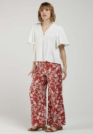 Femme debout portant un chemisier blanc à manches courtes avec un col lacé et un pantalon rouge à jambes larges avec un motif floral blanc et des sandales marron.