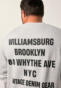 Sudadera gris que presenta un gran texto negro en la parte de atrás: "WILLIAMSBURG BROOKLYN 80 WHYTE AVE NYC VINTAGE DENIM GEAR". Diseño clásico de cuello redondo.