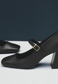 Černé kožené lodičky Mary Jane na vysokém podpatku se čtveratou špičkou a zlatou sponou, vystavené na bílé a tmavě modré ploše.