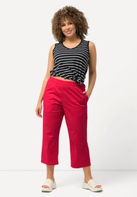 Kvinna med lockigt hår som bär en svart och vit randig ärmlös topp knuten i midjan, ljusröda korta byxor och vita plattformssandaler.