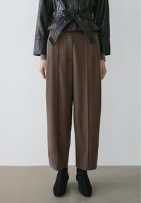 Kvinde iført brune bukser med vide ben, sorte ankelstøvler, grå ærmer og en sort læderjakke med et bundet bælte om taljen.