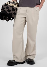 Personne portant un pantalon beige à jambes larges et des chaussures noires épaisses, tenant une veste à carreaux noire et beige avec un lettrage audacieux.