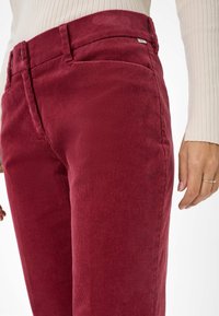 Rode corduroy broek met verticale ribbels, slim fit en zijzakken. Gecombineerd met een crèmekleurig ribbelshirt, met de hand op de heup.
