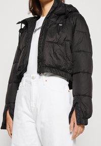 Veste matelassée courte noire avec fermeture éclair à l'avant et capuche. Comprend des poignets élastiques, deux poches avant et une texture brillante et matelassée.