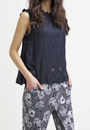 Vrouw draagt een mouwloze marineblauwe geborduurde blouse en zwart-witte bloemetjesbroek met handen in de zakken.