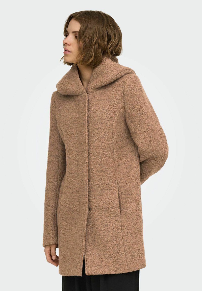 Femme regardant vers la gauche portant un long manteau marron texturé avec un large col et une fermeture frontale cachée.