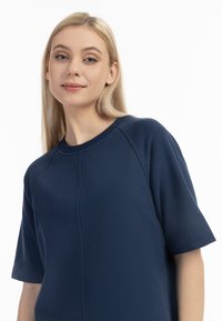 Jonge vrouw met lang blond haar in een marineblauwe T-shirt met korte mouwen, die iets glimlacht, tegen een witte achtergrond.