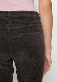 Pantalons en velours côtelé marron avec des côtes verticales, dotés de poches arrière et d'une taille haute. Le tissu a une texture douce et un léger brillant.